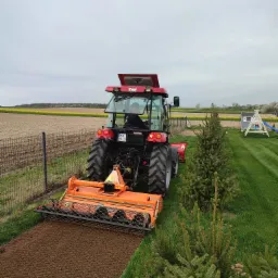 Czerwony traktor z pomarańczowym agregatem uprawowym przygotowuje glebę pod trawnik, widok z tyłu, obok ogrodzenie i iglaki, w tle pole i plac zabaw.