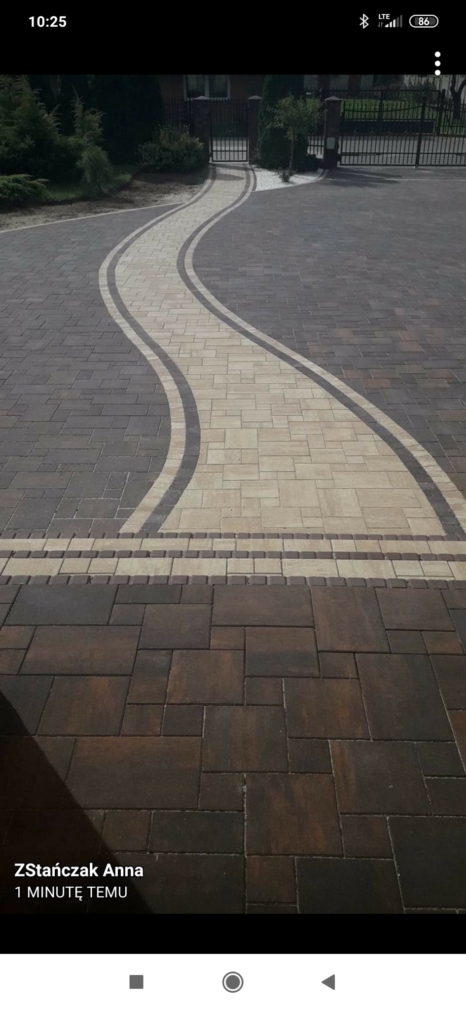 Kręty podjazd z kostki brukowej w dwóch odcieniach szarości i beżu, prowadzący do bramy wjazdowej, z dekoracyjnym obramowaniem z drobnej kostki.