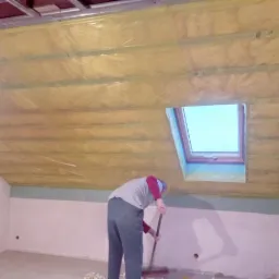 Ocieplenie poddasza wełną mineralną z widocznym oknem dachowym i pracownikiem sprzątającym resztki materiału.