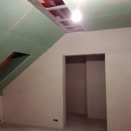 Poddasze w trakcie wykańczania: zamontowane zielone płyty gipsowo-kartonowe na skosach, otwór okienny w płycie, surowy stan ścian, widoczna konstrukcja dachu.