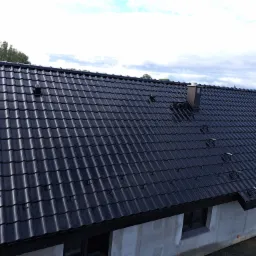 Nowy, błyszczący, czarny dach z dachówki ceramicznej na domu jednorodzinnym, widoczne stopnie kominiarskie i komin. W tle zielony krajobraz.