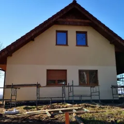 Dwukondygnacyjny dom z beżową elewacją, brązowymi oknami i dachem, otoczony rusztowaniami i widocznym terenem budowy.