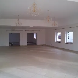 Jasne, przestronne wnętrze z beżową podłogą z płytek, białymi ścianami i kilkoma ozdobnymi żyrandolami. Widoczne okna i otwarte przejścia do innych pomieszczeń.