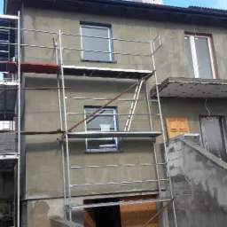 Dwupiętrowy dom w trakcie prac elewacyjnych, z widocznymi rusztowaniami, nowymi oknami zabezpieczonymi taśmą i surową, szarą elewacją przed nałożeniem warstwy wykończeniowej.