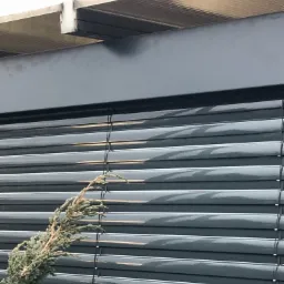 Ciemnoszare żaluzje fasadowe zamontowane na zewnątrz budynku, z widocznym fragmentem rośliny iglastej na pierwszym planie.