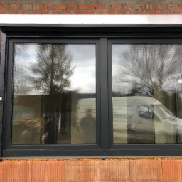 Zamontowane okno PCV w surowej ścianie z cegły, widoczna pianka montażowa, odbicie drzewa i samochodu dostawczego w szybie.