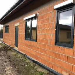 Surowa bryła budynku z czerwonej cegły z zamontowanymi oknami z czarnymi ramami i przygotowanymi otworami na system osłonowy, widoczne rusztowanie i teren po budowie.