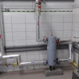 Szare rury instalacji grzewczej z izolacją aluminiową, zaworami i strzałkami wskazującymi kierunek przepływu, zamontowane w pomieszczeniu wyłożonym kafelkami.