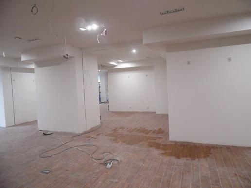 Wnętrze w trakcie remontu: białe ściany, częściowo ułożona podłoga z płytek imitujących drewno, widoczne kable i instalacje elektryczne pod sufitem.