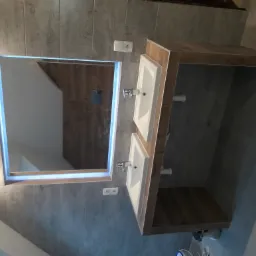 Dwie białe umywalki nablatowe z chromowanymi bateriami, osadzone na drewnopodobnym blacie w nowoczesnej łazience z szarymi płytkami imitującymi beton, podświetlane lustro LED i widoczne gniazdka...