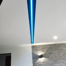 Nowoczesny sufit z podłużnym panelem LED emitującym niebieskie światło, kontrastujący z białą powierzchnią sufitu i dekoracyjną ścianą z kamienia w odcieniach szarości z zamontowanymi kinkietami.