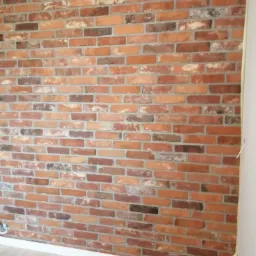 Ściana z cegły klinkierowej w odcieniach czerwieni i brązu, z szarą fugą, wykończona przy podłodze białą listwą przypodłogową, widoczne zabezpieczenie taśmą malarską na styku ze ścianą boczną.