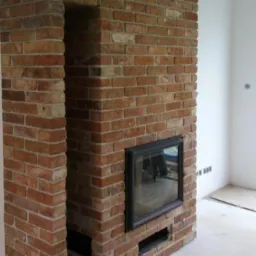 Murowany kominek z cegły w surowym wnętrzu, z widocznymi otworami wentylacyjnymi i czarną ramą paleniska, na tle białych ścian i surowej posadzki.