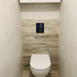 Nowoczesna toaleta podwieszana z białym sedesem i chromowanym przyciskiem spłukującym na tle płytek imitujących drewno, otoczona białymi płytkami ściennymi.