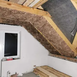 Poddasze z izolacją z wełny mineralnej pomiędzy krokwiami, widoczne deski stropowe, okno dachowe i przygotowane deski na podłodze.