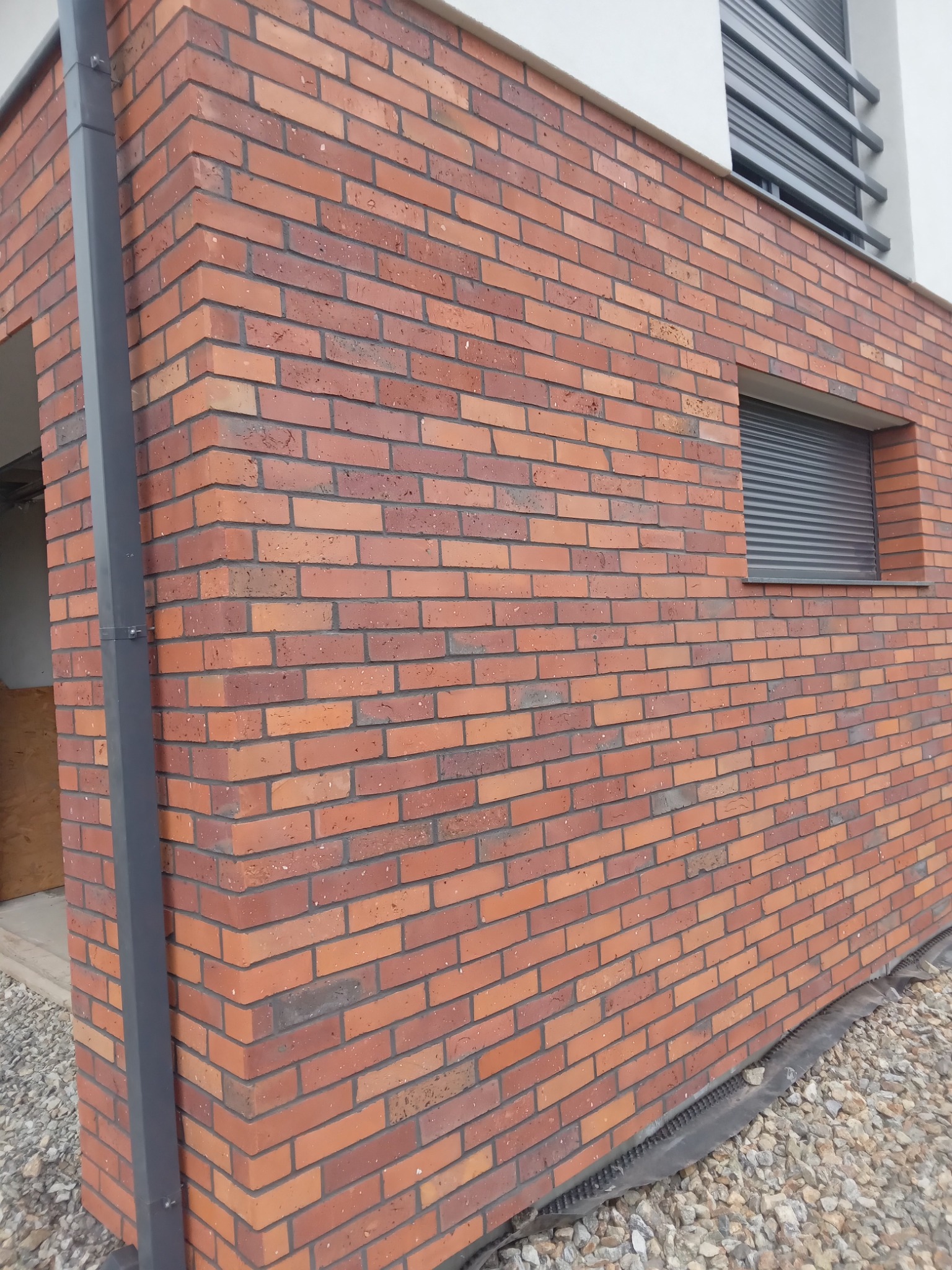 Fragment elewacji budynku z czerwonej cegły klinkierowej, widoczny narożnik, okno z roletą zewnętrzną i rynna spustowa; wokół budynku rozsypany żwir.