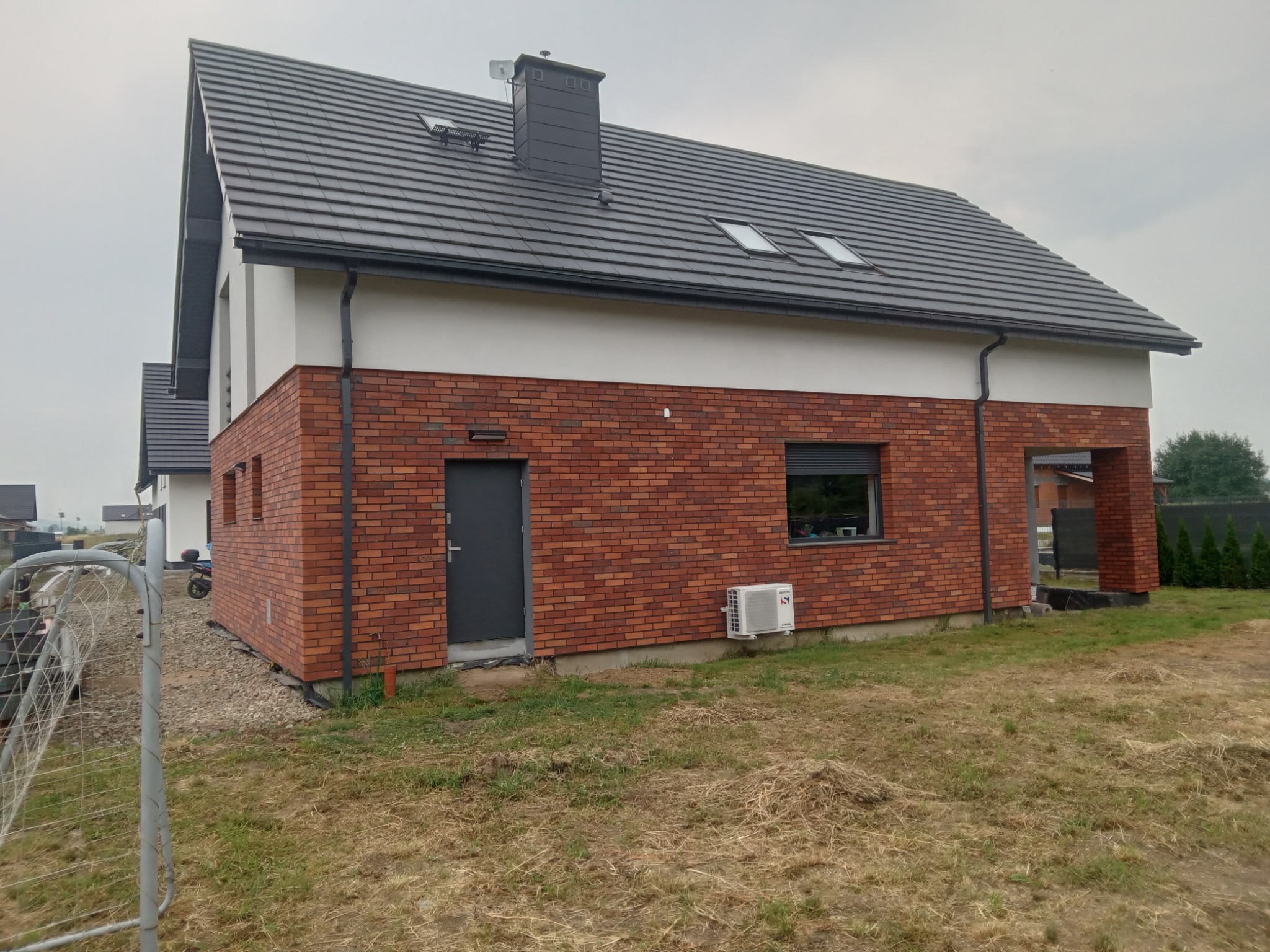 Nowoczesny dom jednorodzinny z czerwonej cegły klinkierowej, szarą dachówką i czarnymi elementami wykończeniowymi, widoczny z zewnątrz w pochmurny dzień.