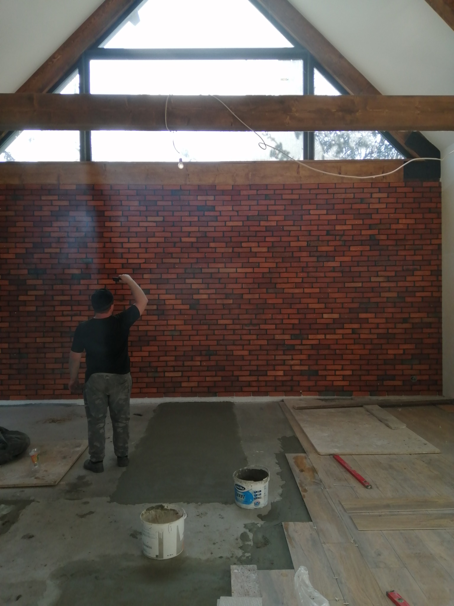 Montaż cegieł dekoracyjnych na ścianie w pomieszczeniu z drewnianą konstrukcją stropu, widoczne narzędzia i materiały budowlane na podłodze.