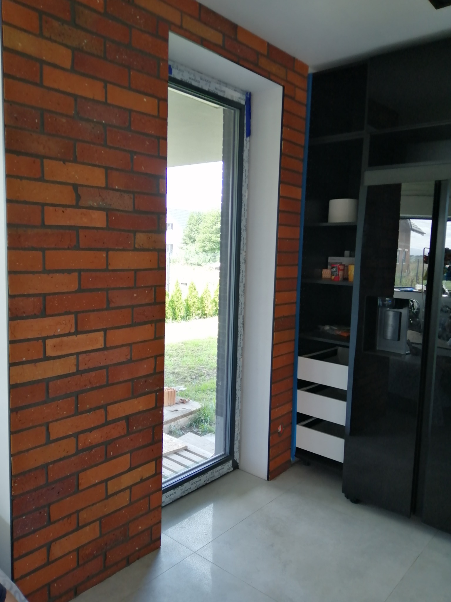 Ściana z cegły klinkierowej obok okna z widokiem na ogród, przylegająca do czarnej zabudowy kuchennej z białymi szufladami, na jasnej podłodze.