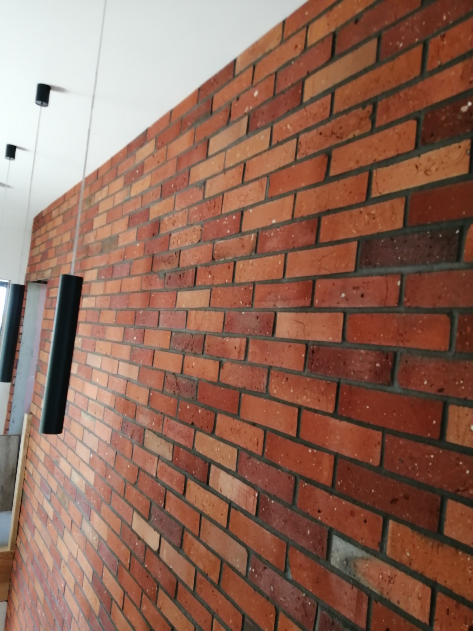 Ściana wykończona czerwoną cegłą klinkierową z szarą fugą, oświetlona wiszącymi, czarnymi lampami tubami.