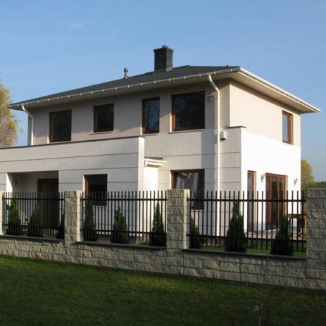 Dwukondygnacyjny dom z jasną elewacją, czarnym dachem i ogrodzeniem z kamienia łupanego oraz metalowych sztachet, na tle zielonego trawnika i błękitnego nieba.