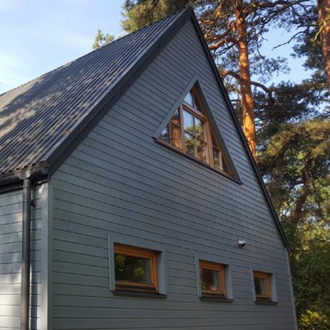 Szary dom z drewnianą elewacją, trójkątnym oknem na szczycie i małymi kwadratowymi oknami na dole, otoczony drzewami.