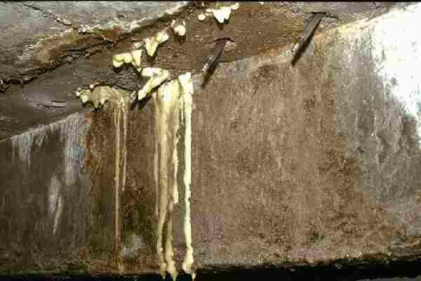 Nieszczelny betonowy strop z widocznymi prętami zbrojeniowymi i wyciekającą pianą poliuretanową, prawdopodobnie używaną do uszczelnienia