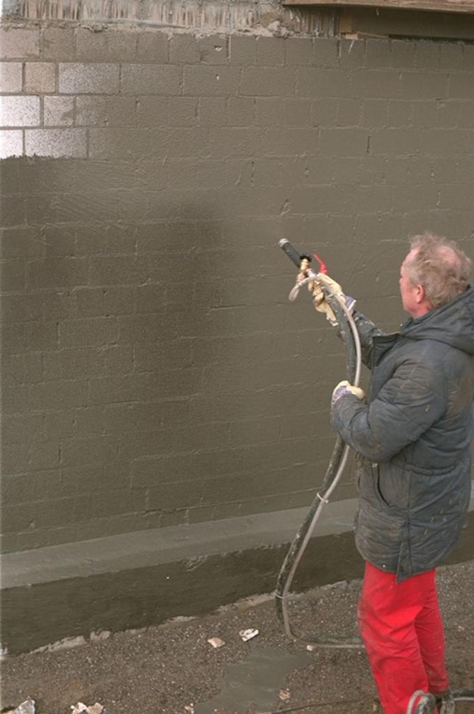 Pracownik w czerwonych spodniach natryskuje szlam bitumiczno-akrylowy na ścianę z bloczków betonowych, tworząc hydroizolację.