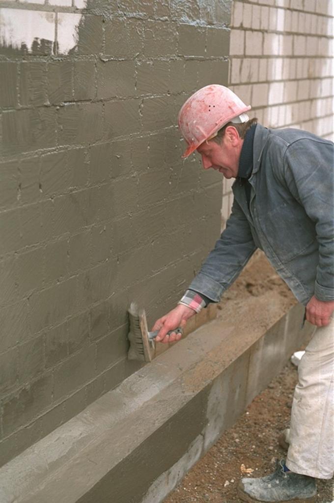 Pracownik w kasku maluje szlamem bitumiczno-akrylowym ścianę fundamentową z bloczków betonowych, używając pędzla dekarskiego.