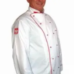 Mężczyzna w białym fartuchu kucharskim z czerwoną lamówką i czapce kucharskiej, z naszywką z flagą Polski na ramieniu.