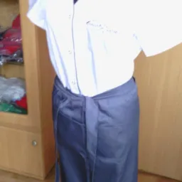 Fartuch wiązany w pasie, biała bluza z krótkim rękawem i szarym wykończeniem kołnierza na manekinie, widoczny fragment szafy w tle.