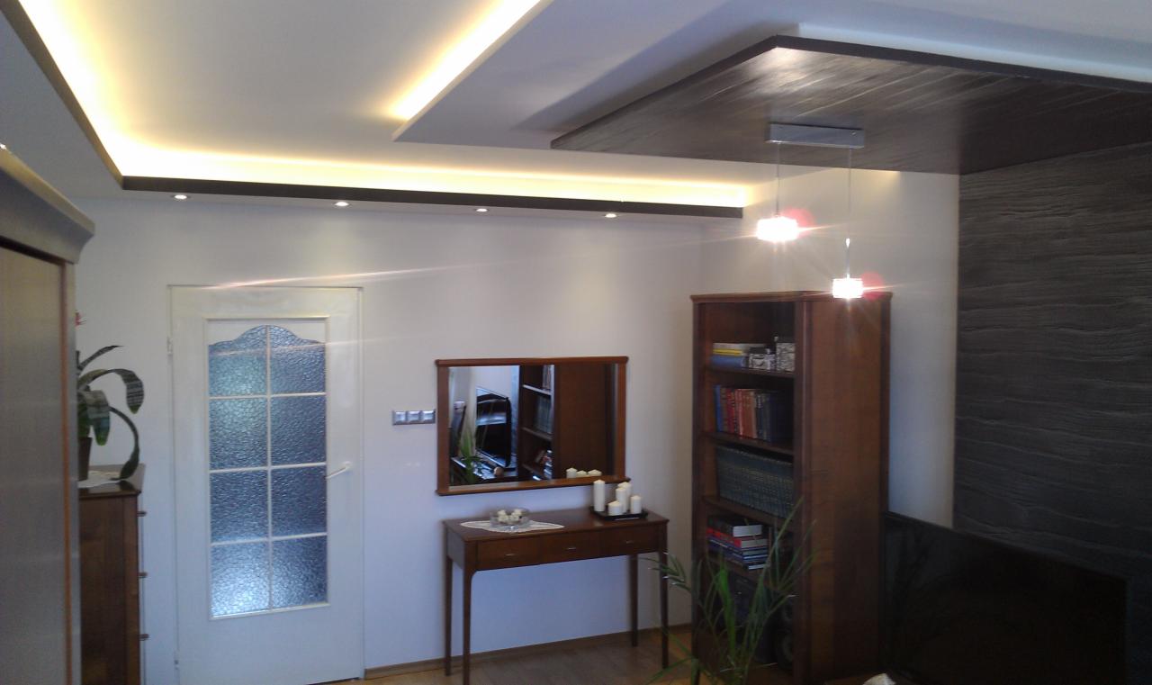 Nowoczesny salon z podwieszanym sufitem z oświetleniem LED, drewnianą boazerią ścienną i lustrem nad konsolą. Widoczna zabudowa meblowa i drzwi z szybą.