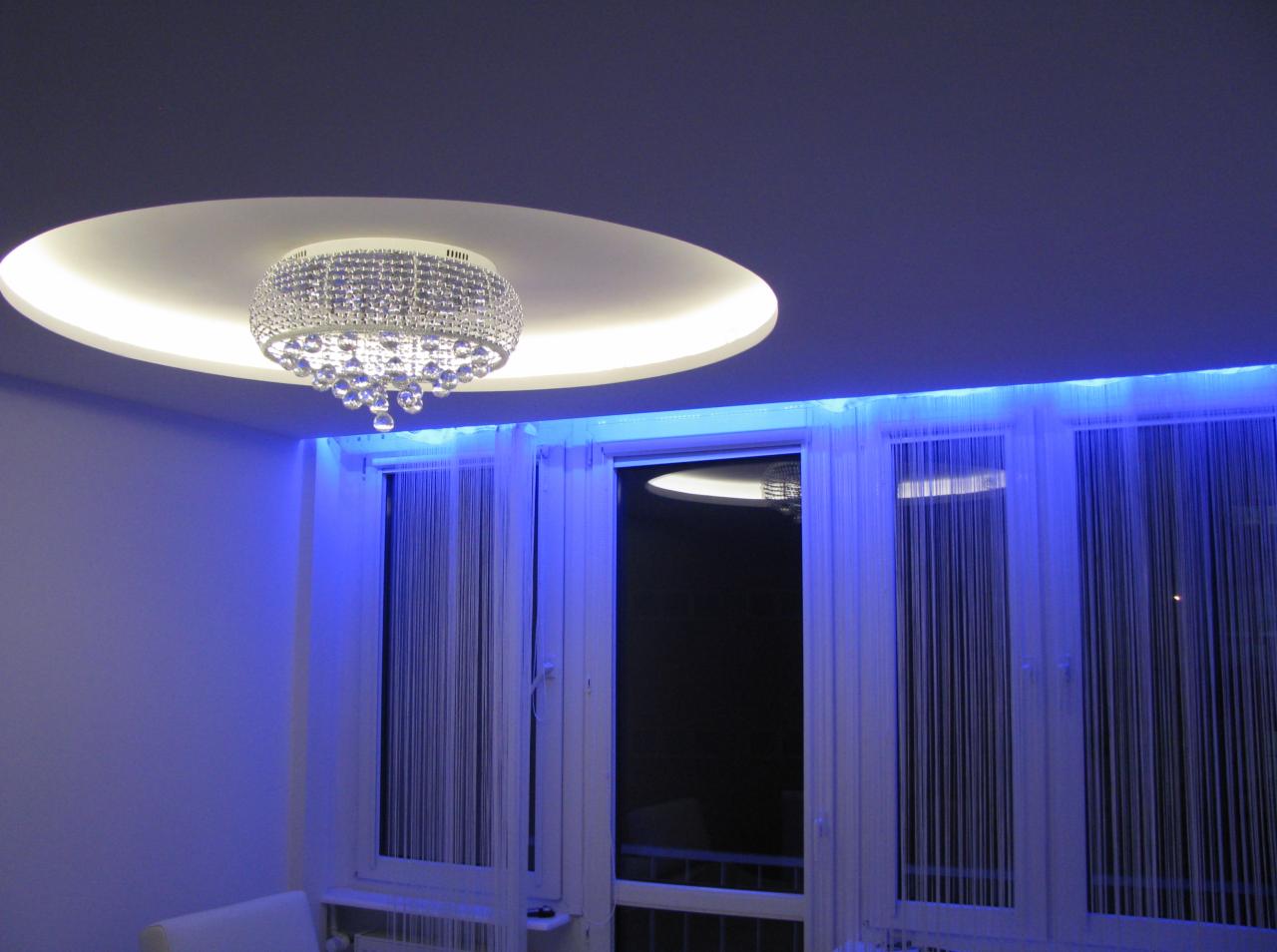 Owalny sufit podwieszany z oświetleniem LED i kryształowym żyrandolem. Okna z białymi ramami i sznurkowymi zasłonami, podświetlone niebieskim światłem.