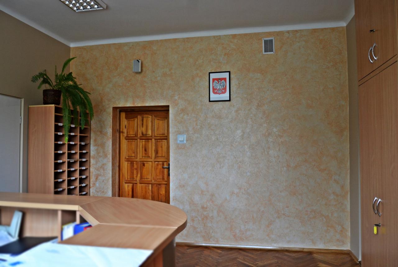 Wykończona ściana z efektem dekoracyjnym w odcieniach beżu, drewniane drzwi i parkiet w jodełkę. Na ścianie wisi godło Polski w czarnej ramce.