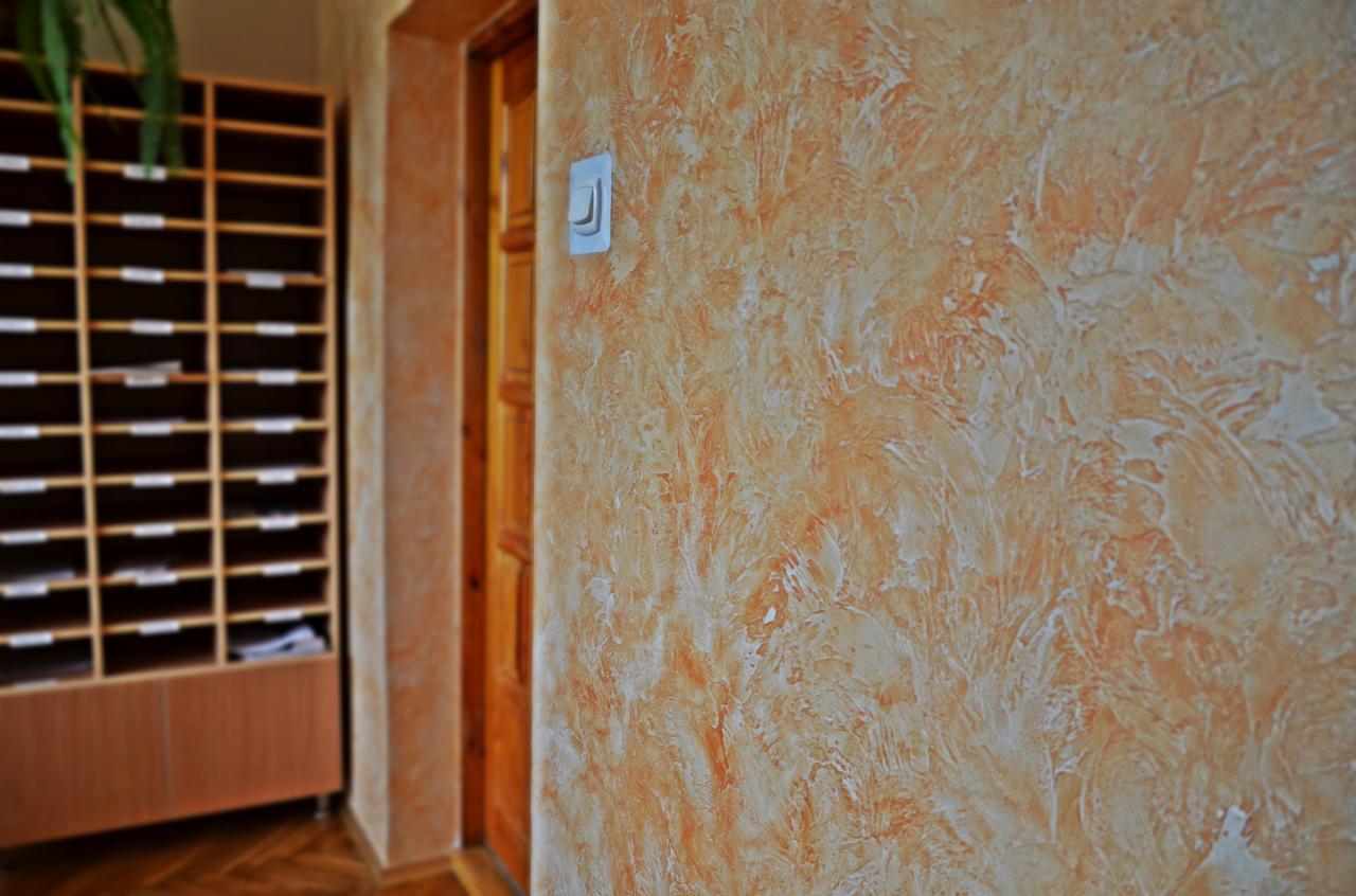 Ściana z dekoracyjnym tynkiem w odcieniach pomarańczy i bieli, widoczny włącznik światła i fragment drewnianych drzwi oraz regał na dokumenty w tle.