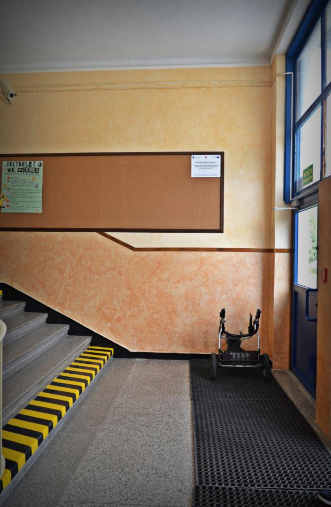 Korytarz szkolny z marmurkową ścianą w odcieniach beżu i pomarańczy. Schody z żółto-czarnymi pasami, tablica korkowa i wózek inwalidzki przy drzwiach.