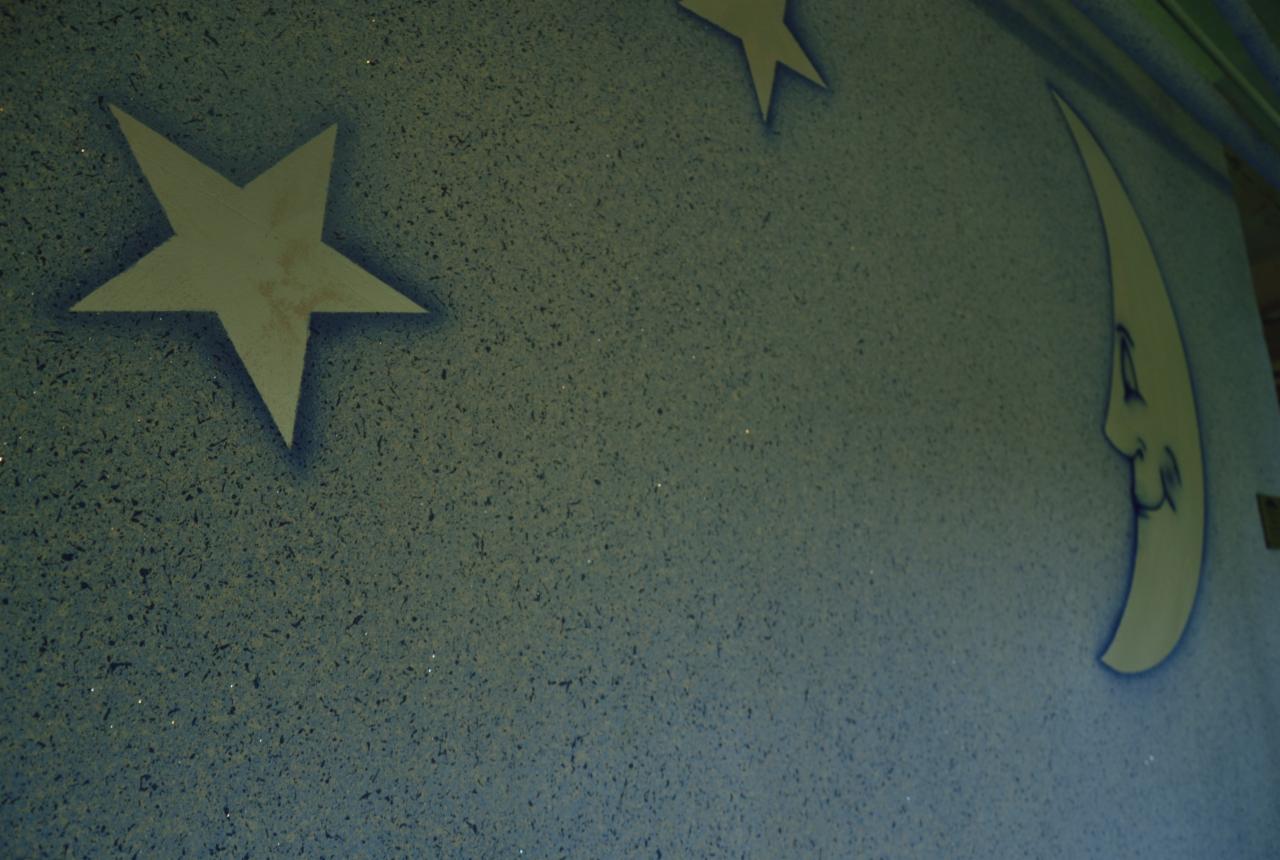 Niebieska ściana z dekoracyjnym malowaniem: gwiazdy i księżyc z twarzą, wykonane techniką natryskową. Drobna faktura tła, delikatne cieniowanie.