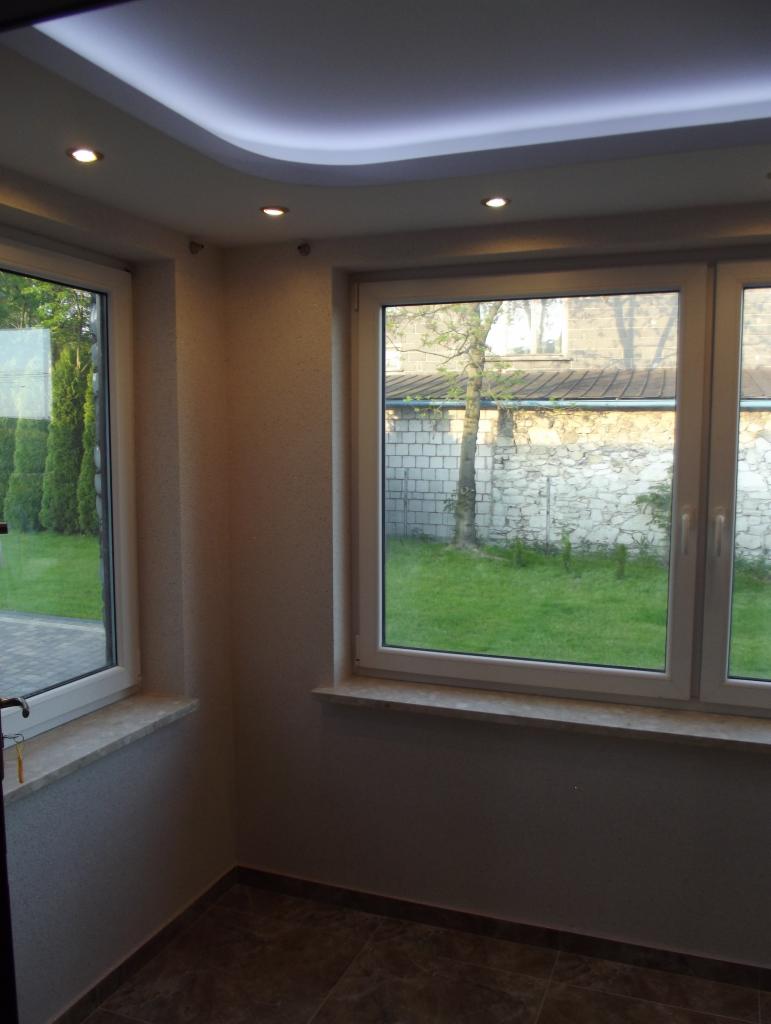 Wykończone wnętrze z oknami na ogród, podwieszany sufit z oświetleniem punktowym i brązową podłogą. Widok na zewnątrz przez okna.