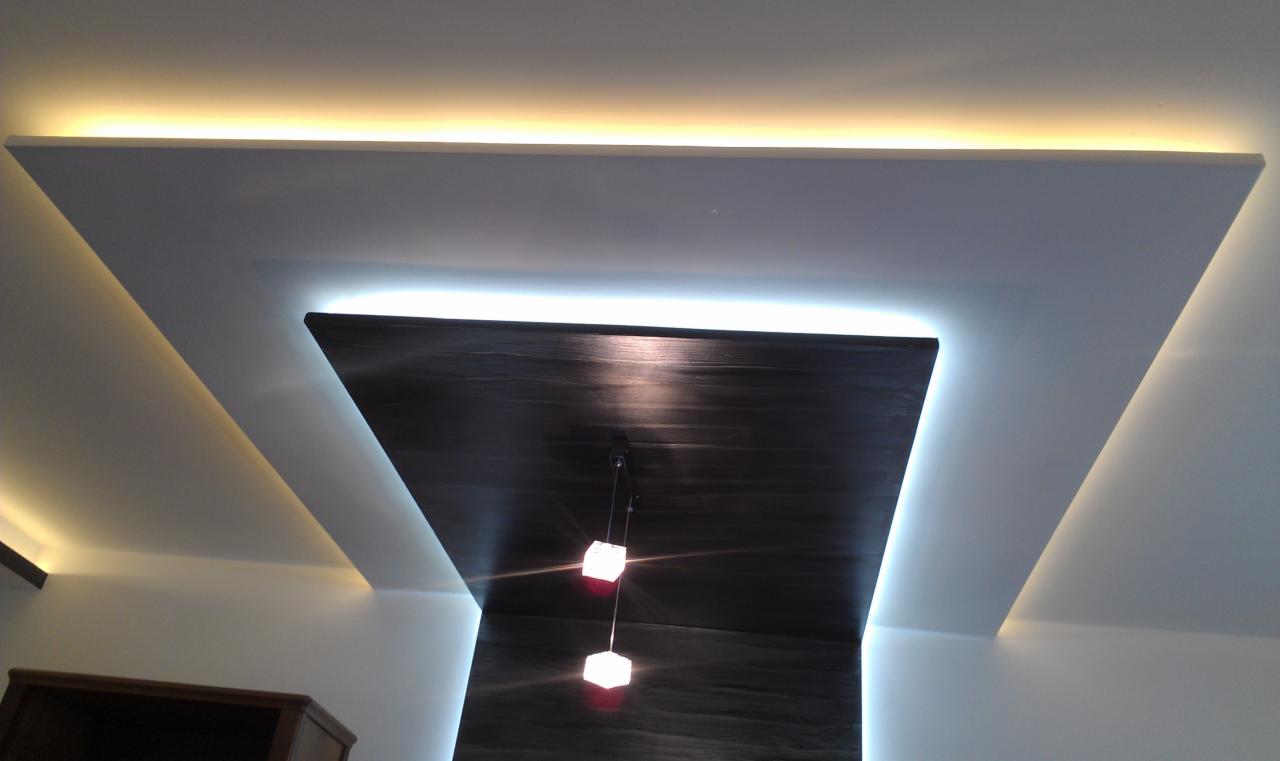 Podwieszany sufit z płyt karton-gips, oświetlenie LED tworzące geometryczne wzory, drewniana wstawka i minimalistyczne lampy wiszące. Nowoczesny design.