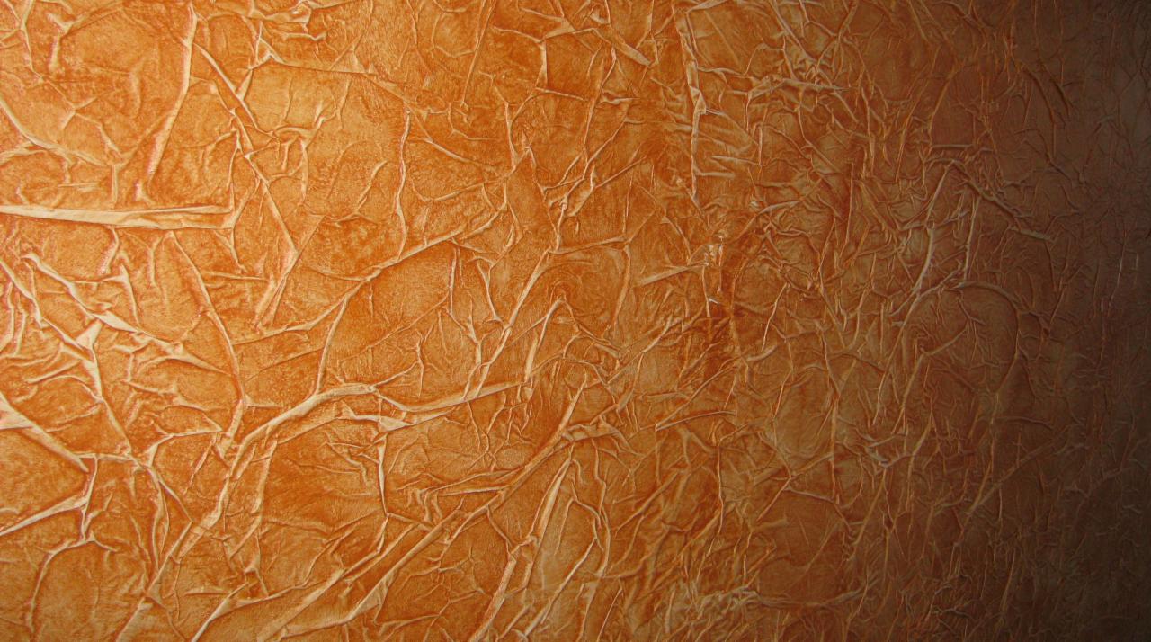 Pomarańczowa ściana z wyraźną, nieregularną fakturą przypominającą popękany tynk. Cieniowanie od jasnego do ciemnego podkreśla trójwymiarowość wzoru.