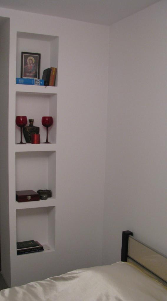 Biała ściana z wnękami, w których znajdują się dekoracyjne przedmioty: książki, ikona, kieliszki i butelka. Widoczny fragment łóżka.