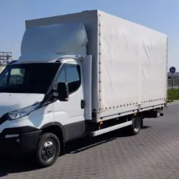 Biały samochód dostawczy Iveco z plandeką na tle przemysłowego krajobrazu i czerwonych flag reklamowych.