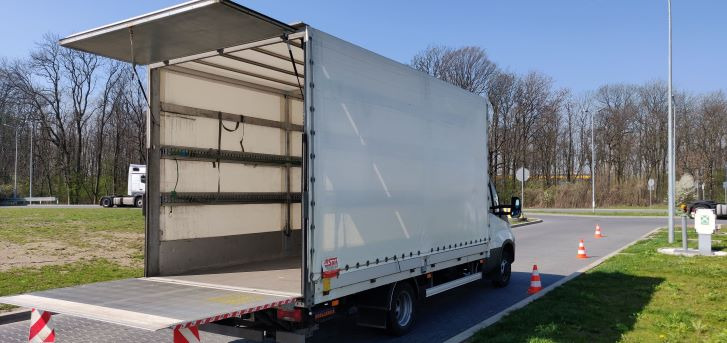 Otwarty, biały samochód dostawczy z podniesioną klapą, widoczne metalowe mocowania wewnątrz, zaparkowany na parkingu z pomarańczowymi pachołkami.