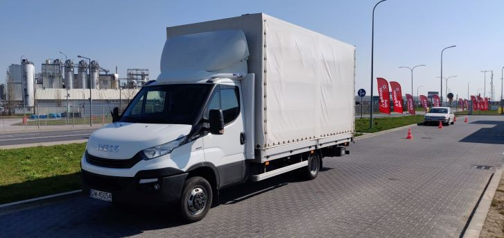 Biały samochód dostawczy Iveco z plandeką na tle przemysłowego krajobrazu i czerwonych flag reklamowych.