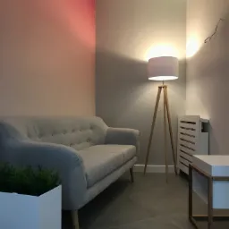 Jasnoszara sofa z guzikami, drewniany stolik kawowy z białą górą i donica z zielenią, oświetlone przez lampę stojącą na trójnogu w minimalistycznym wnętrzu z akcentem kolorystycznym na ścianie.