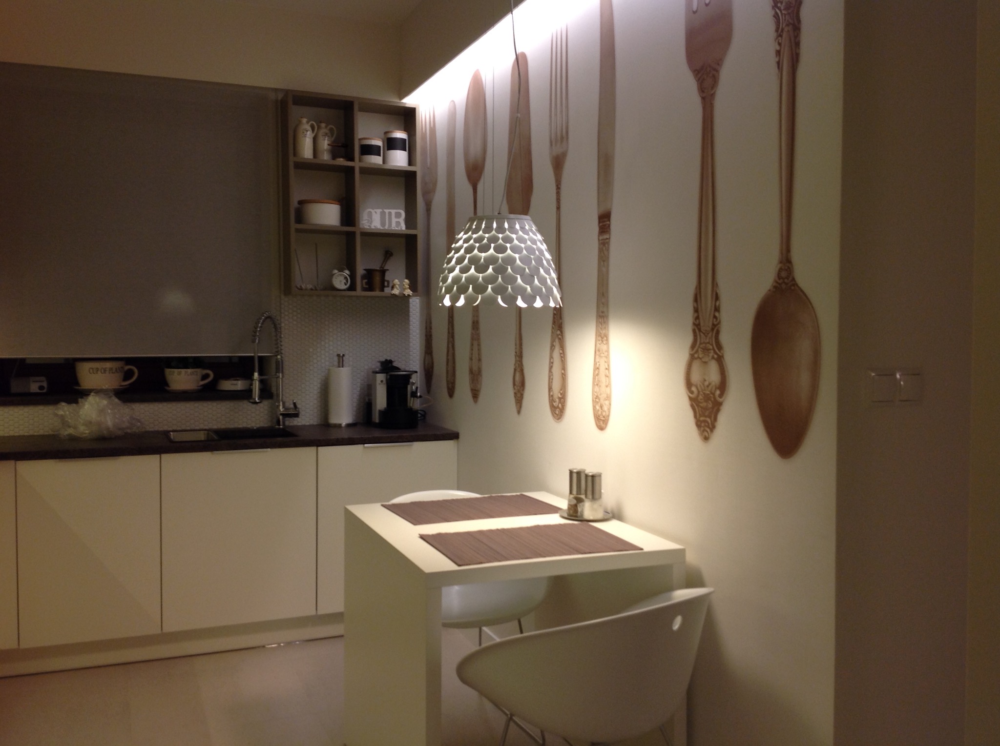 Nowoczesna kuchnia z białymi szafkami, małym stołem z dwoma krzesłami i dekoracyjną tapetą z motywem sztućców, oświetlona designerską lampą.