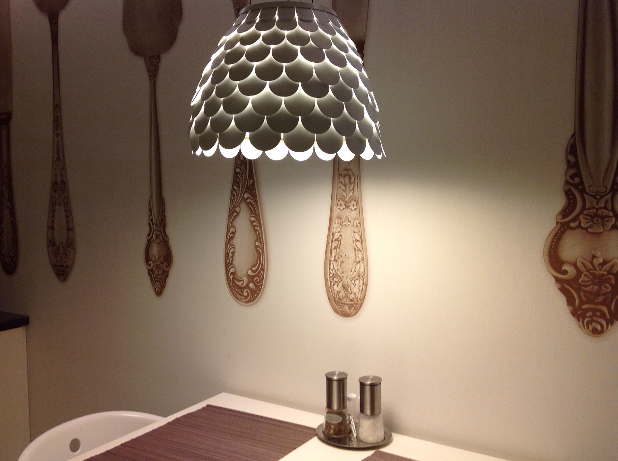 Jadalnia z dekoracyjną lampą w kształcie łuski i ozdobami ściennymi imitującymi sztućce, solniczka i pieprzniczka na białym stole.