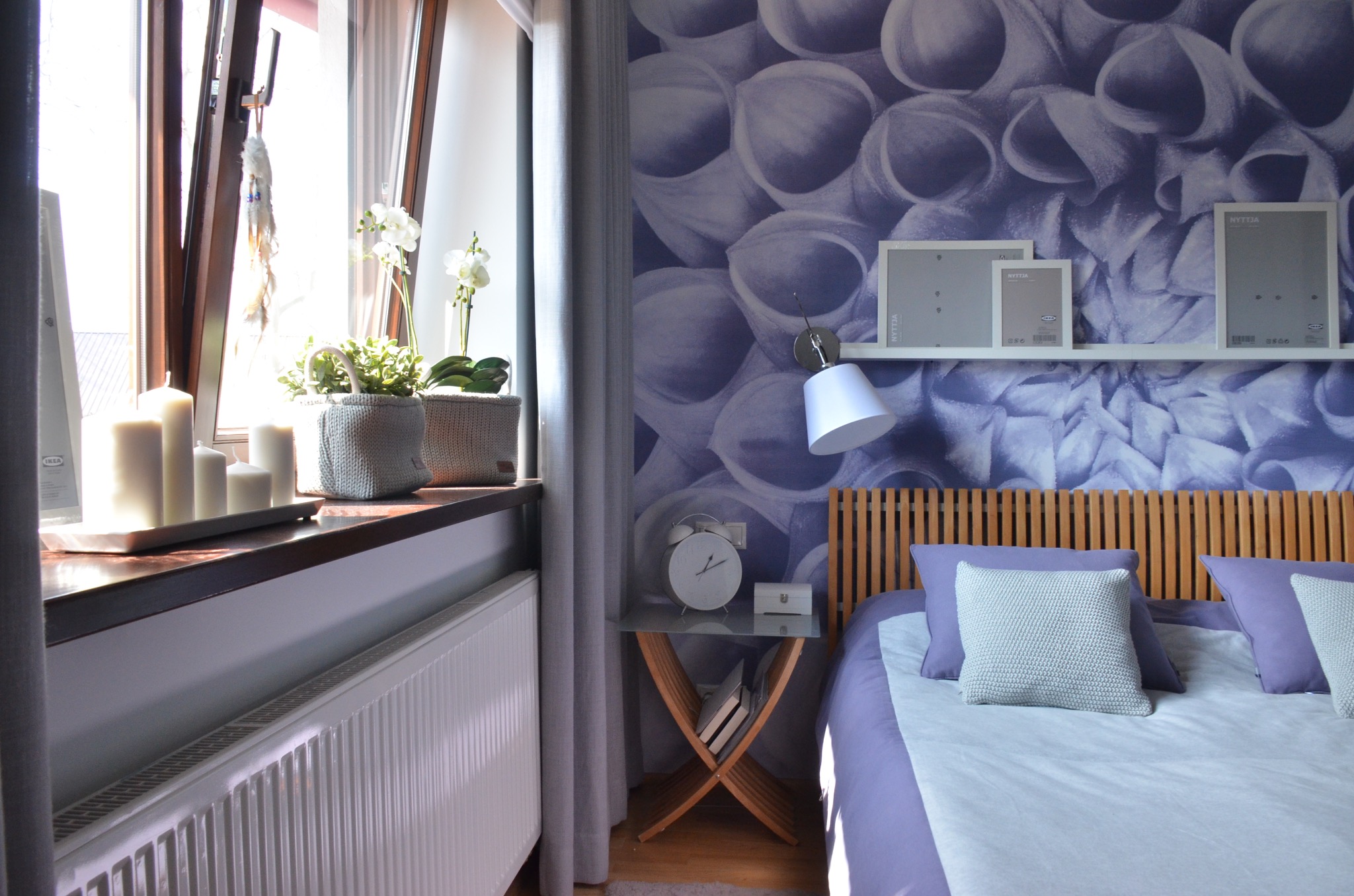 Jasna sypialnia z drewnianym zagłówkiem, fioletowymi akcentami i tapetą w kwiaty, stolik nocny z zegarem i książkami, okno z roślinami i świecami.