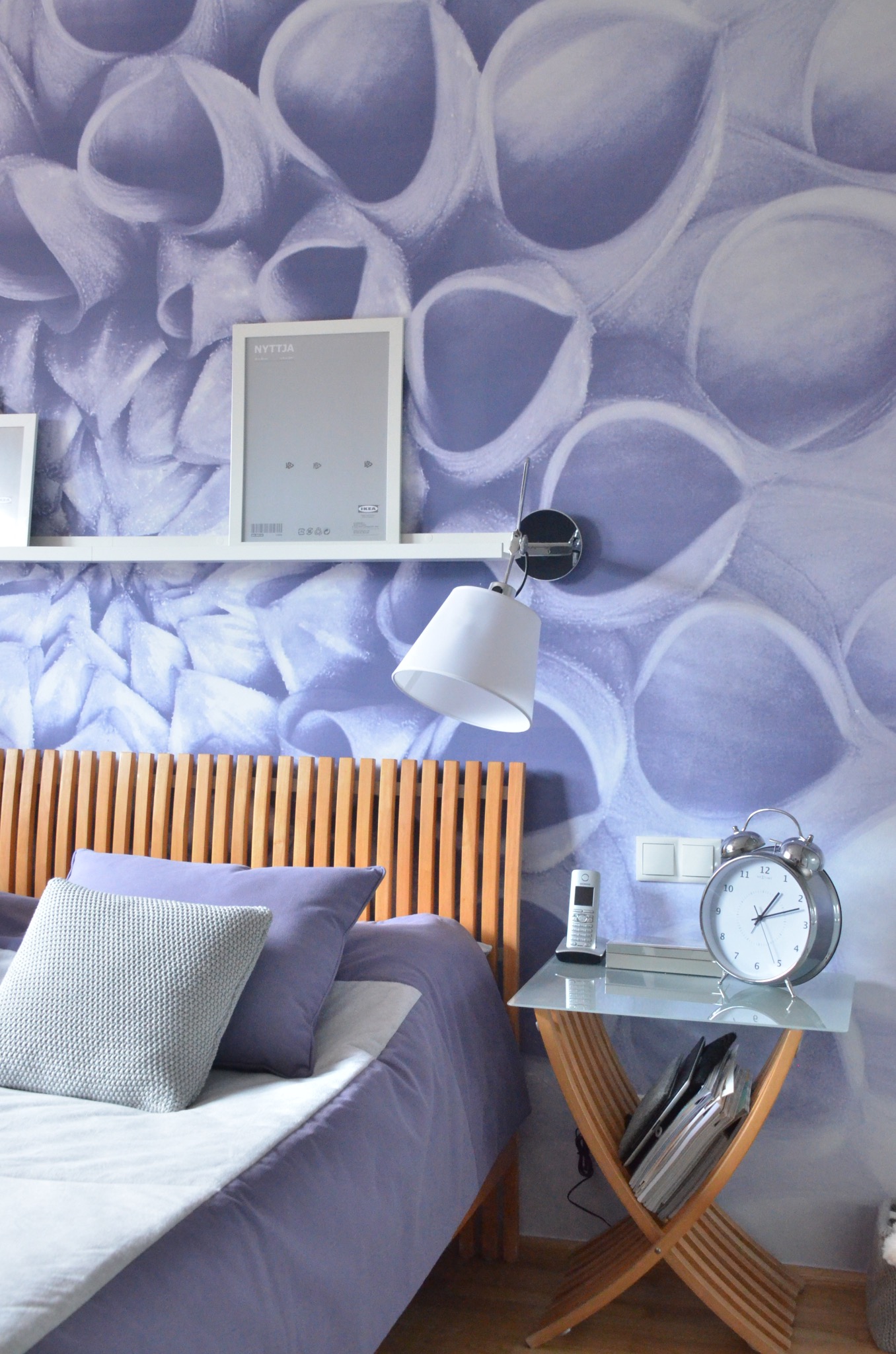 Sypialnia z drewnianym wezgłowiem łóżka, fioletową pościelą, tapetą w abstrakcyjne wzory, półką z ramkami, lampką nocną i stolikiem z budzikiem.