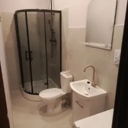 Nowoczesna łazienka po remoncie: kabina prysznicowa z czarnymi profilami, biała umywalka z szafką i lustro na tle beżowych płytek. Minimalistyczny design.